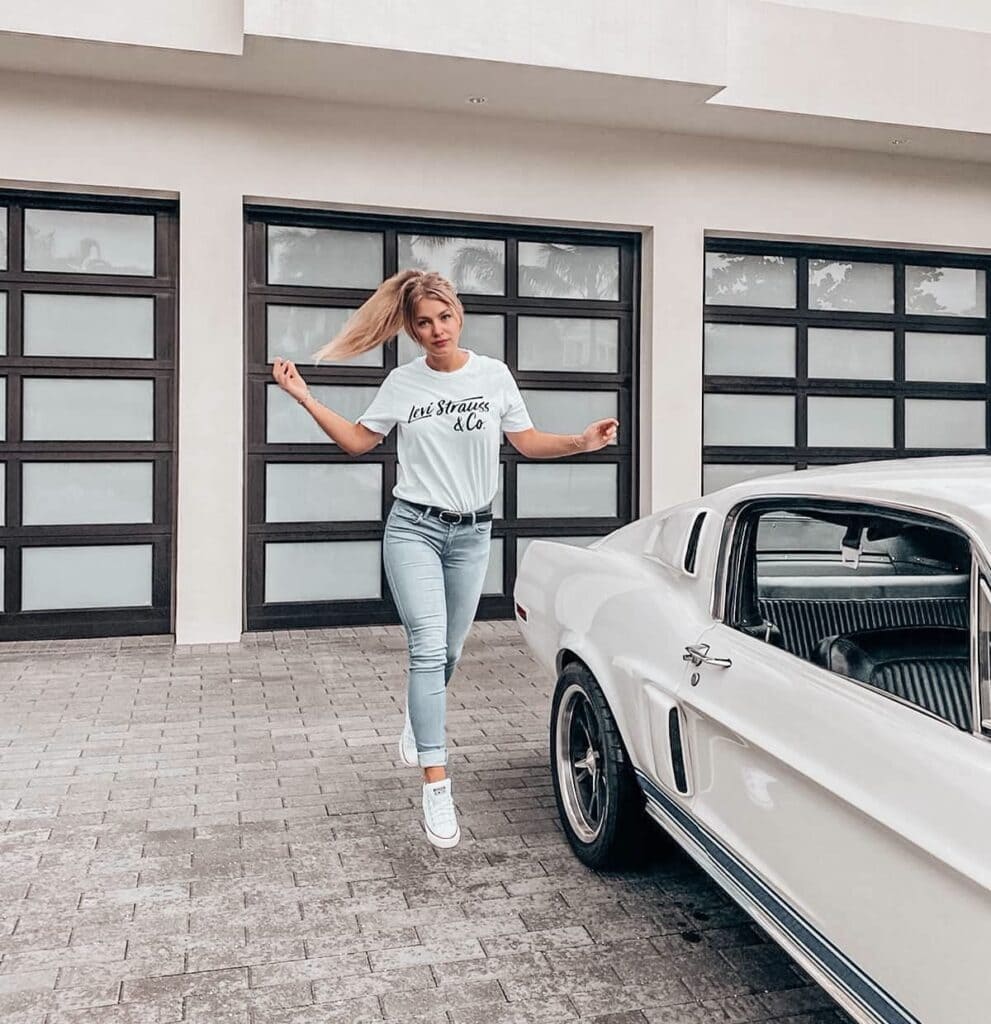 Elsie Graham in jeans and a white t - shirt is standing next to a white car.